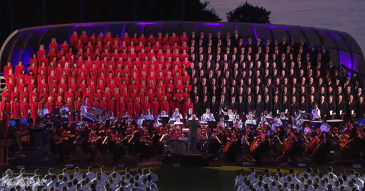 A choir performance at an outdoor concert venue with a stage, orchestra, conductor, and audience under purple stage lights; trees are visible in the background indicating dusk or dawn.