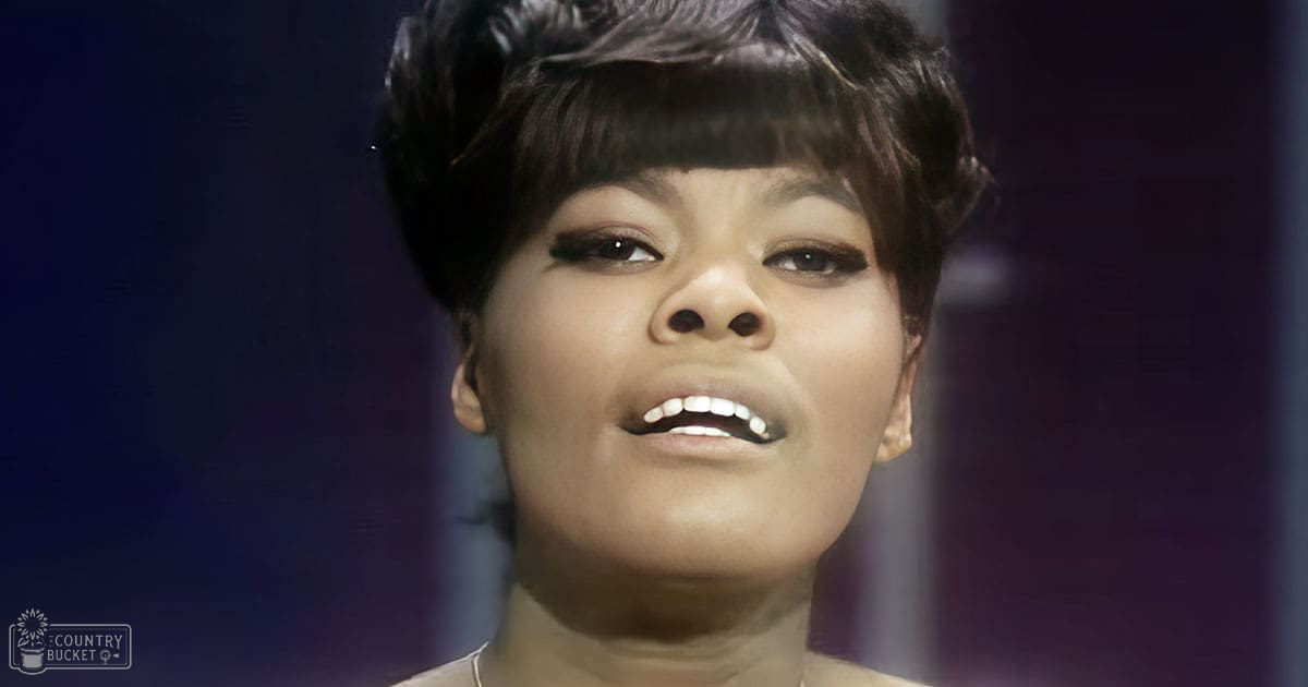 Young adult female wearing an elegant, dark-colored garment with a high neckline and short sleeves, likely mid-20th century style, indoors with dim lighting and dark purples and blues dominating the background.