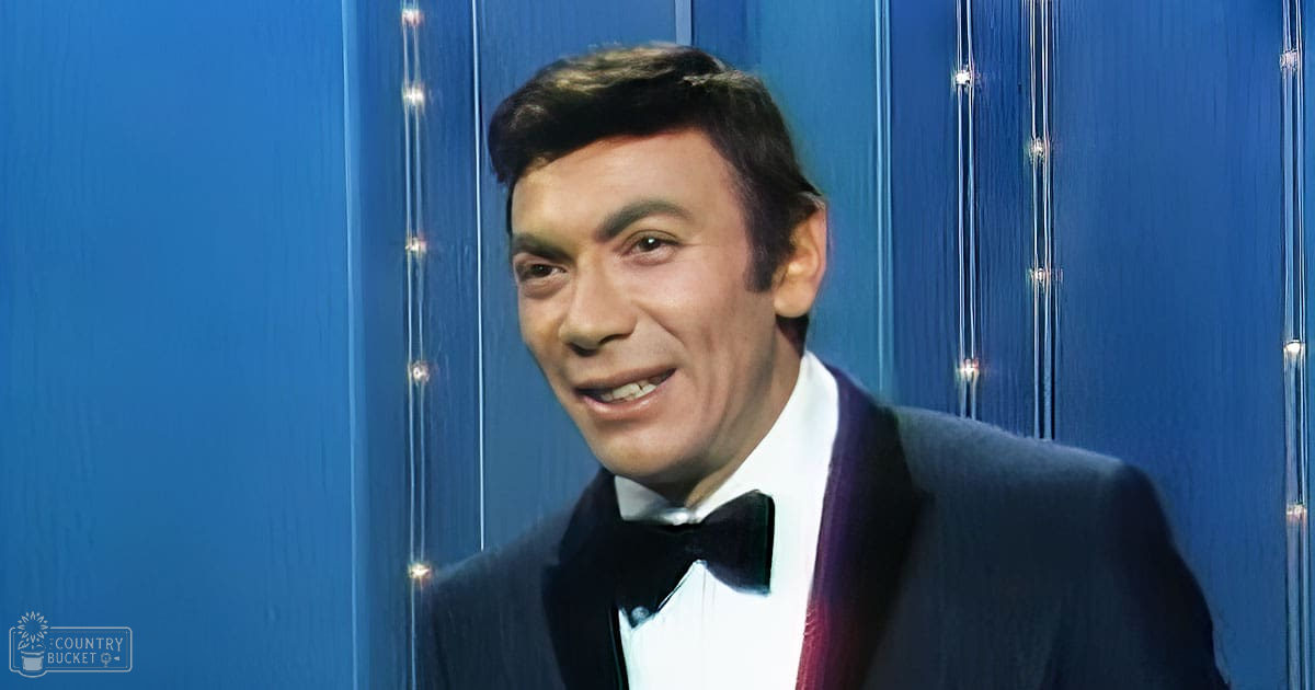 An adult male in a formal black tuxedo with bow tie on stage or studio setting.
