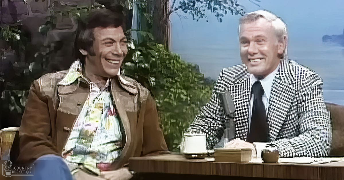 Two individuals seated at a desk on a talk show set with indoor studio lighting and decorative plants in the background.
