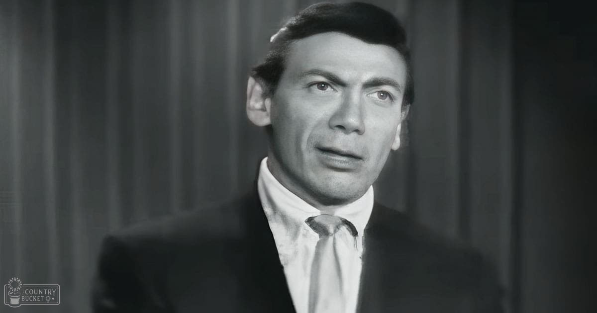 A middle-aged man wearing a dark suit jacket, white shirt, and light-colored tie in an indoor setting with curtains as part of the background.