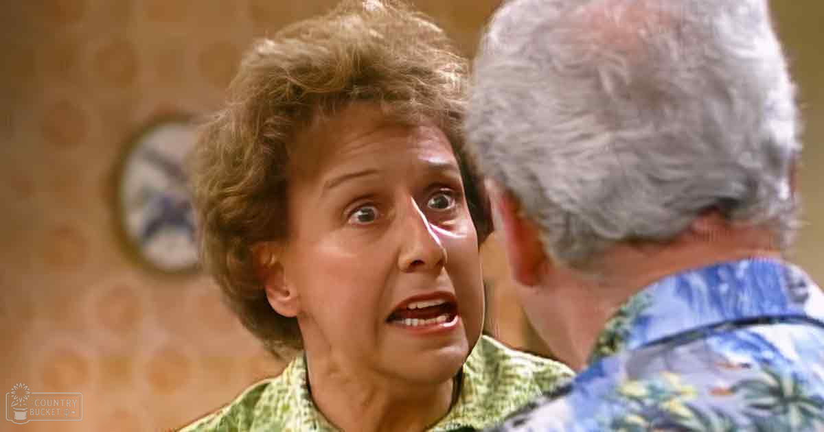 Middle-aged woman in a green patterned shirt indoors with an older adult man wearing a blue Hawaiian-style shirt nearby; indoor setting, artificial lighting, patterned wallpaper and clock visible.