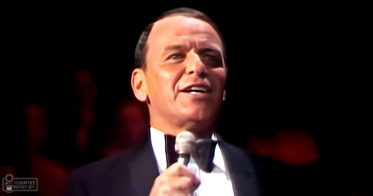 An adult male performer in a dark suit jacket with a white shirt, holding a microphone on stage under dim lighting.