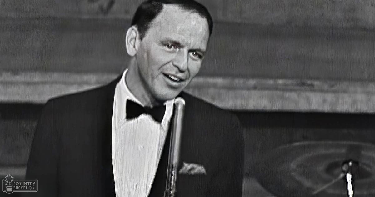 A middle-aged man in a formal tuxedo with a bow tie stands on an indoor stage or studio, surrounded by a microphone and part of a drum set.