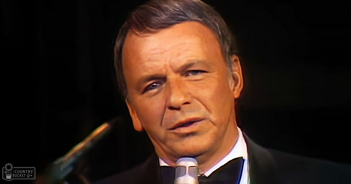 A middle-aged adult male wearing a dark suit jacket and white collar shirt, standing indoors with stage lighting.