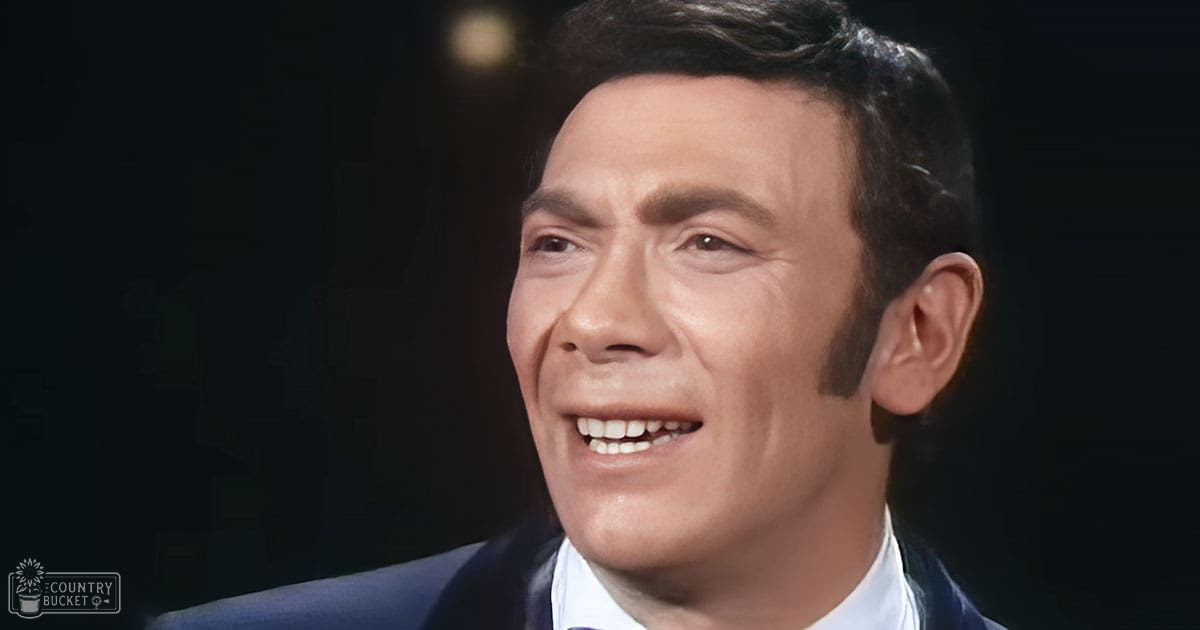 An adult male in a dark blue suit and white shirt, standing against a dark backdrop with soft lighting, likely in a studio setting.