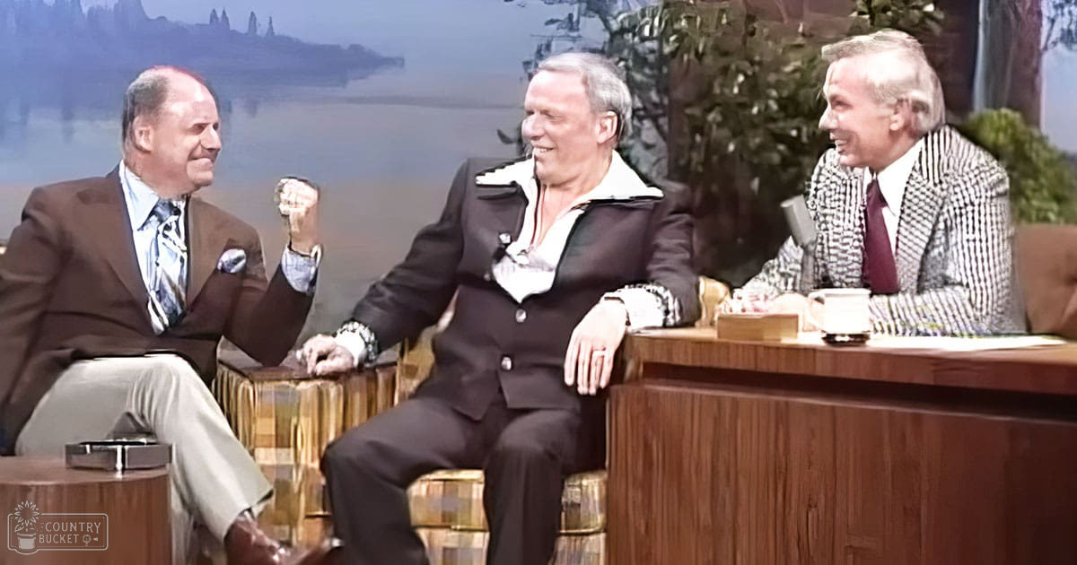 Three adult males in a studio environment discussing on a talk show set against a scenic backdrop of water and mountains or trees.