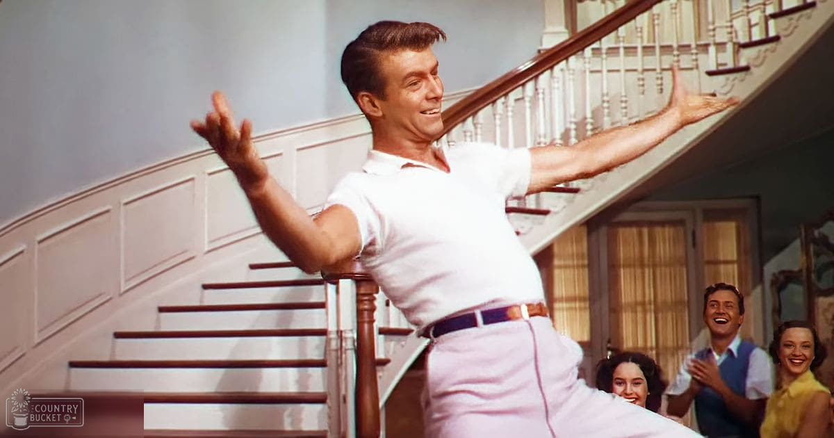 An adult male in a white polo shirt and light pink pants stands indoors, possibly dancing or performing; background includes two adults clapping, one in a blue vest over a white shirt and the other in a yellow dress.