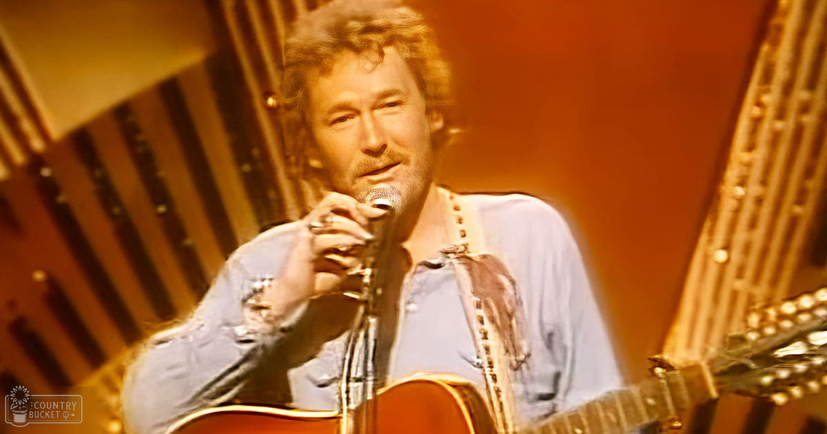 A middle-aged adult male wearing a denim shirt over rolled-up sleeves, holding a microphone while standing on what appears to be a stage or performance area with musical instruments and warm lighting.