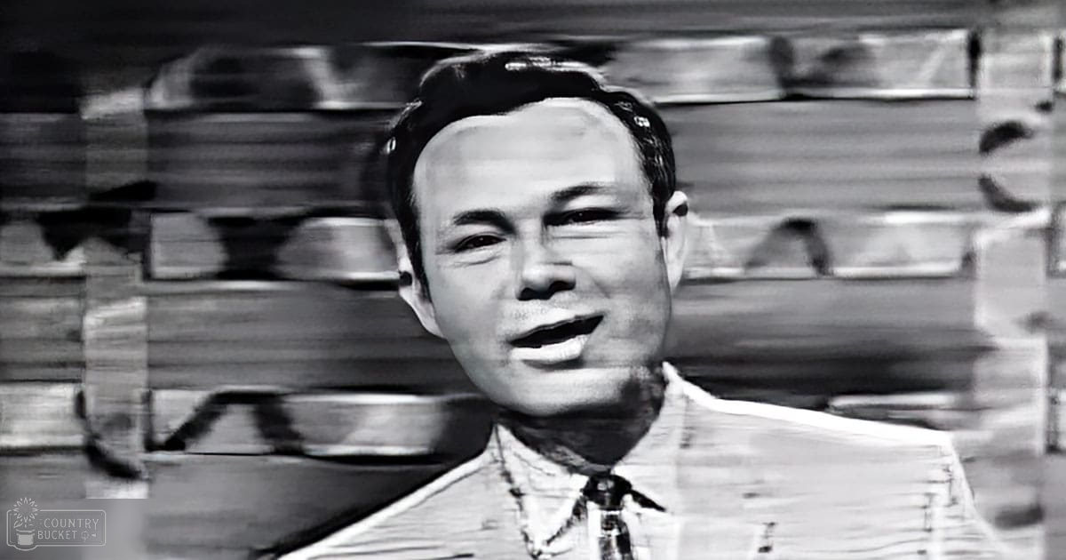 A young adult male in an indoor setting, wearing a patterned shirt with a tie and a collared jacket or blazer over it.