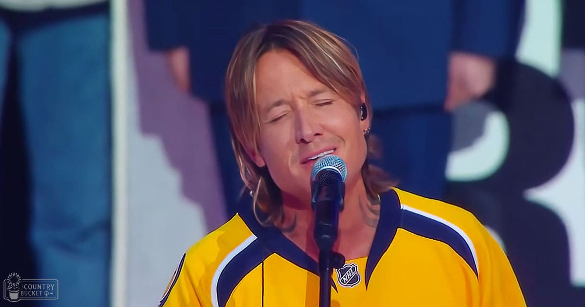 Adult male performer wearing a yellow and blue NHL hockey jersey, singing into a microphone at what appears to be an indoor event in front of a hockey arena.