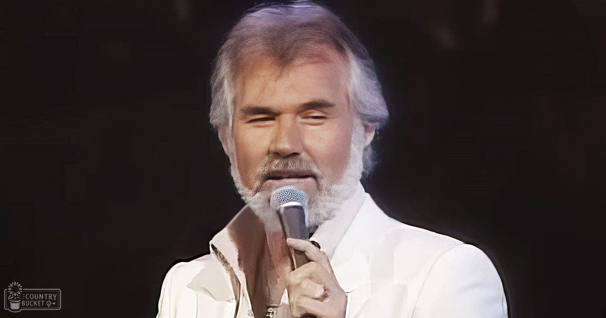 A middle-aged adult male wearing a white, relaxed shirt with long sleeves slightly rolled up, holding a microphone on stage under dark spotlighting, suggesting an indoor performance setting.