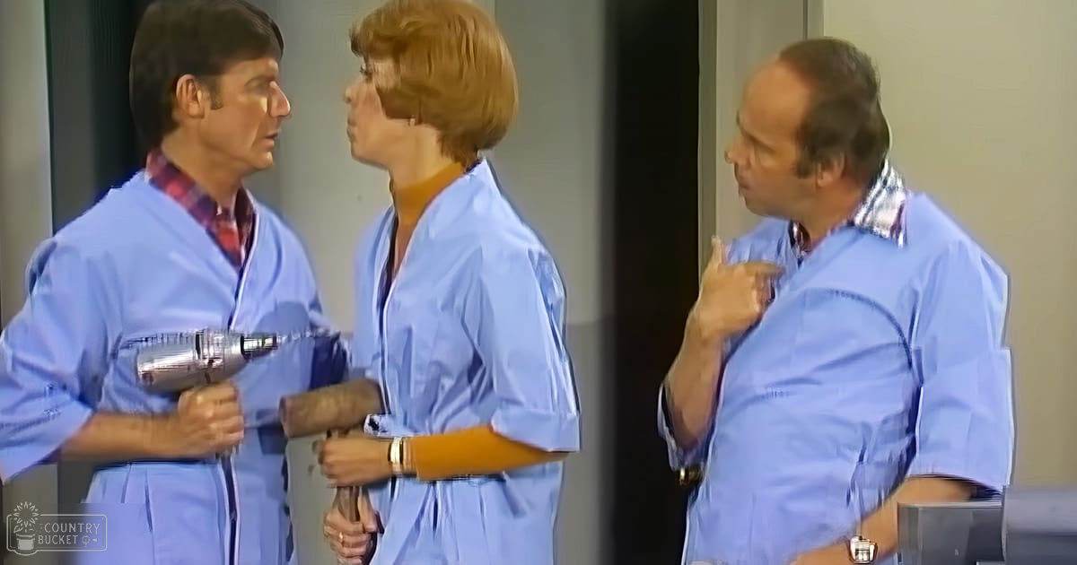 Three individuals wearing similar light blue robes with white trim, interacting in a simple indoor setting with minimal decor.