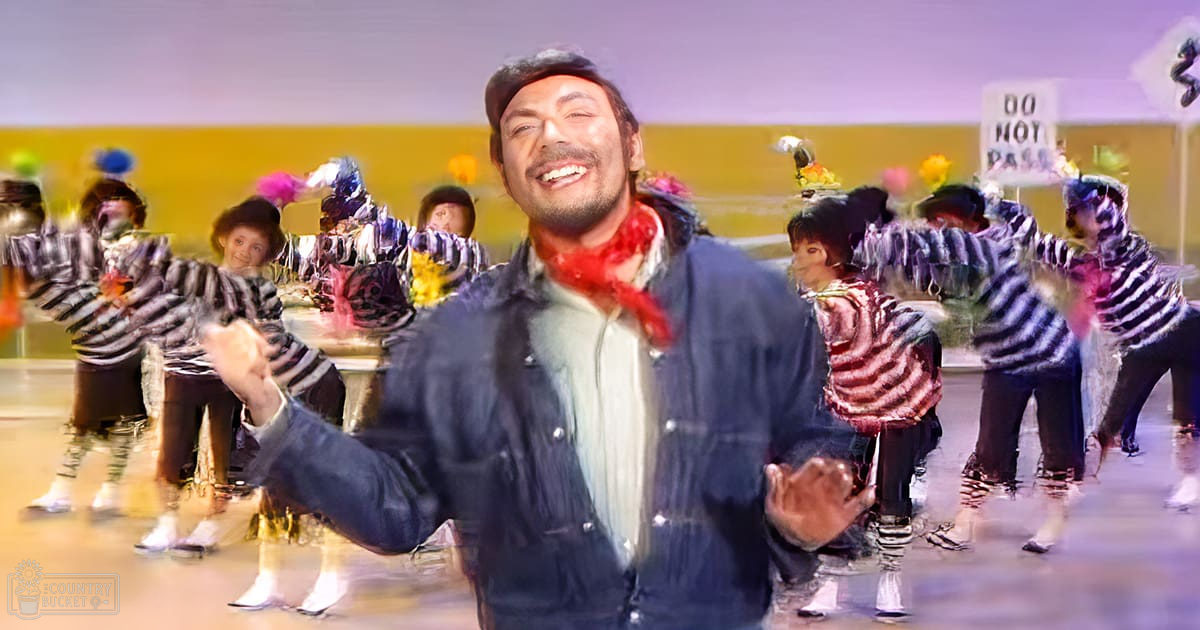 A young adult male dancing in front of a group of children dressed in striped shirts indoors.