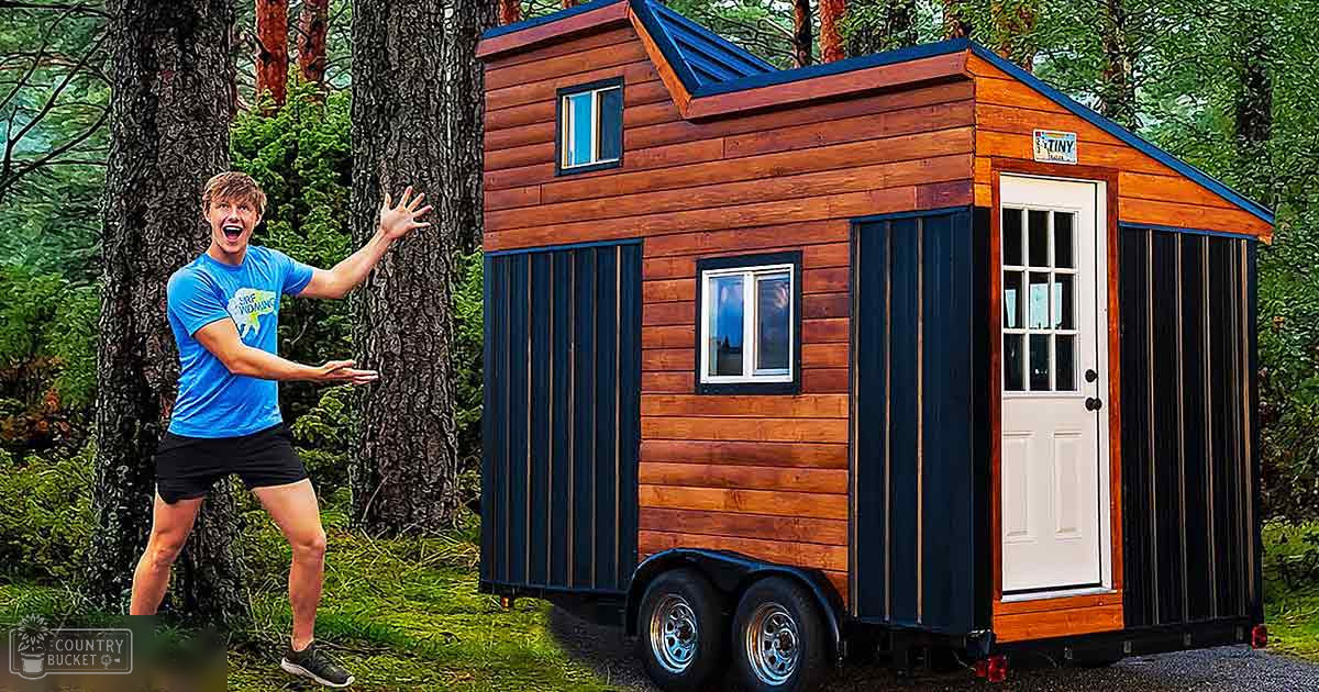 A young adult male wearing a blue 'Surf Wyoming' t-shirt, black shorts, and black athletic shoes stands in a forested area with a small wooden structure resembling a tiny house or trailer.