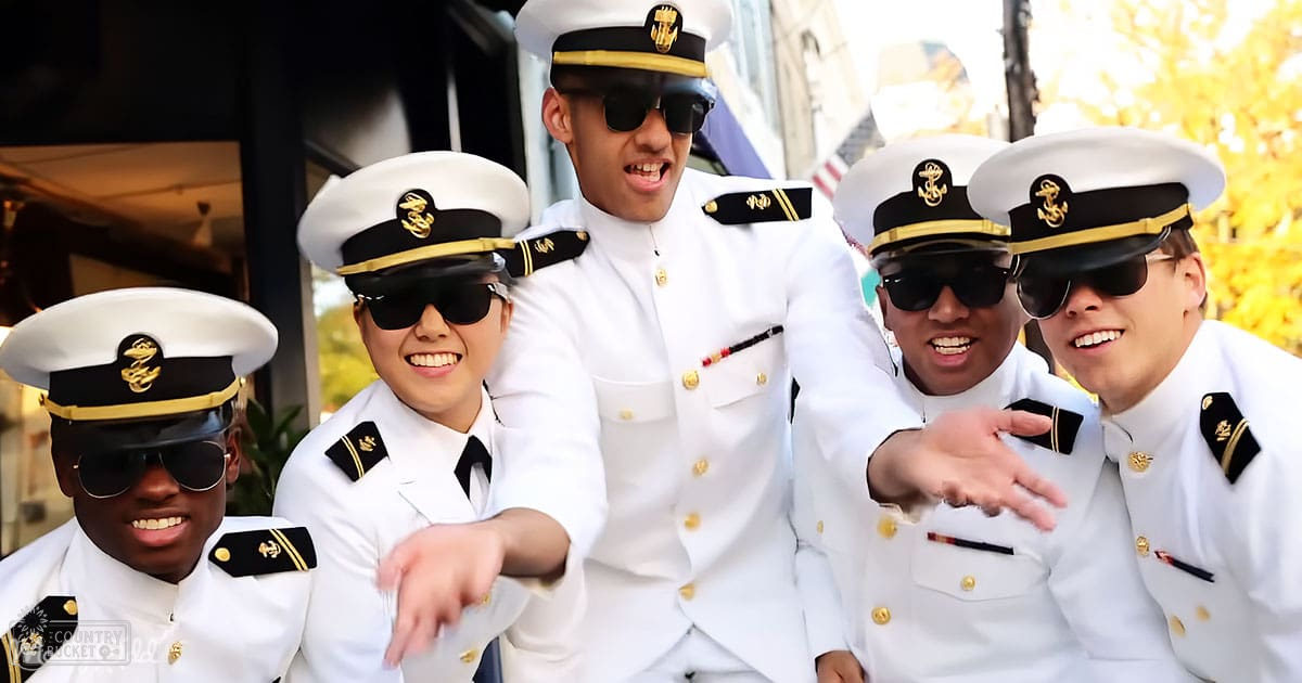 A group of young adults in white naval-style uniforms with gold buttons and black trims are gathered outdoors near a building with large windows, surrounded by greenery. The scene suggests an event or gathering during autumn daytime.