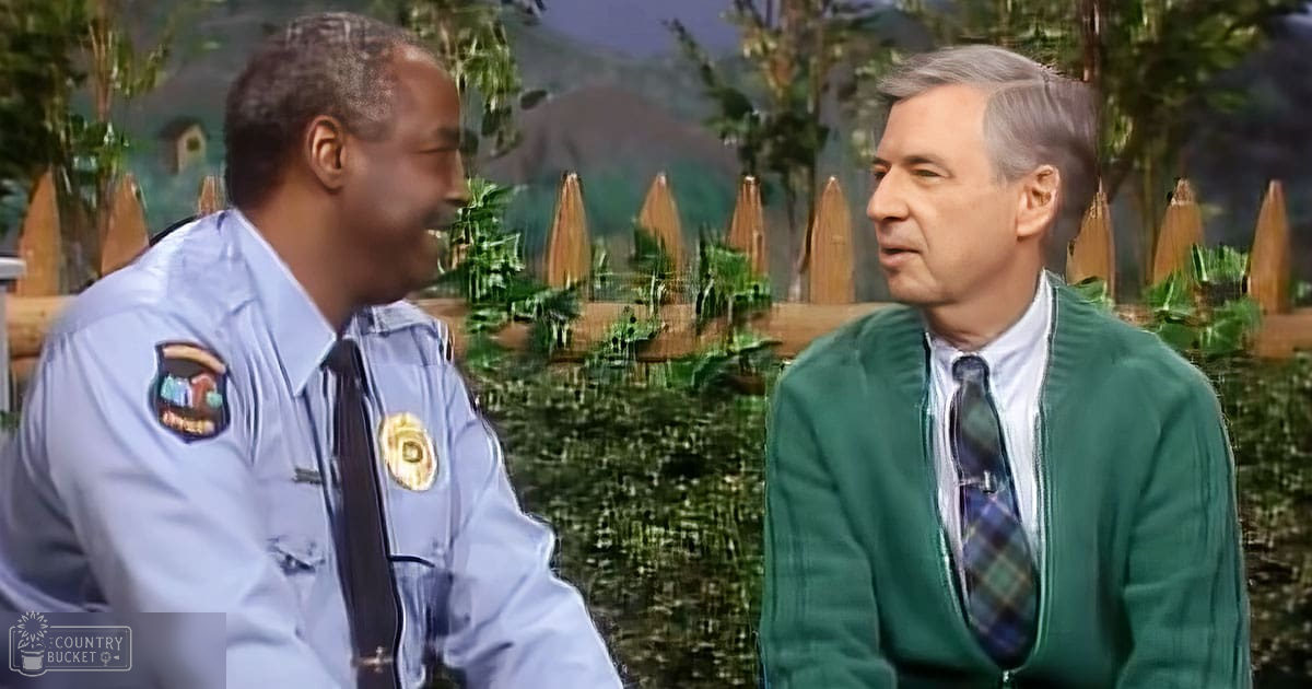 An outdoor television or movie set with a male adult in a blue uniform and another middle-aged to older adult man in formal attire, surrounded by natural scenery during daytime.
