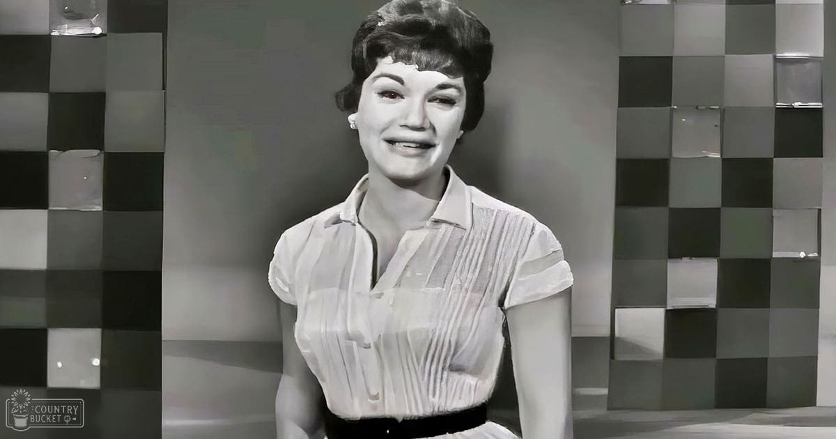 A young adult female stands against a checkered wall, dressed in a white pleated blouse and dark pants, under even frontal studio lighting.