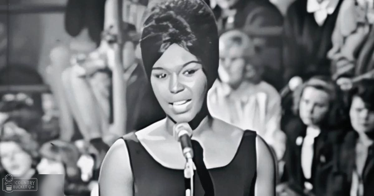 A young adult female singer wearing a sleeveless dress with a high neckline performs into a microphone in front of an audience dressed in formal attire at what appears to be a stage or performance venue.
