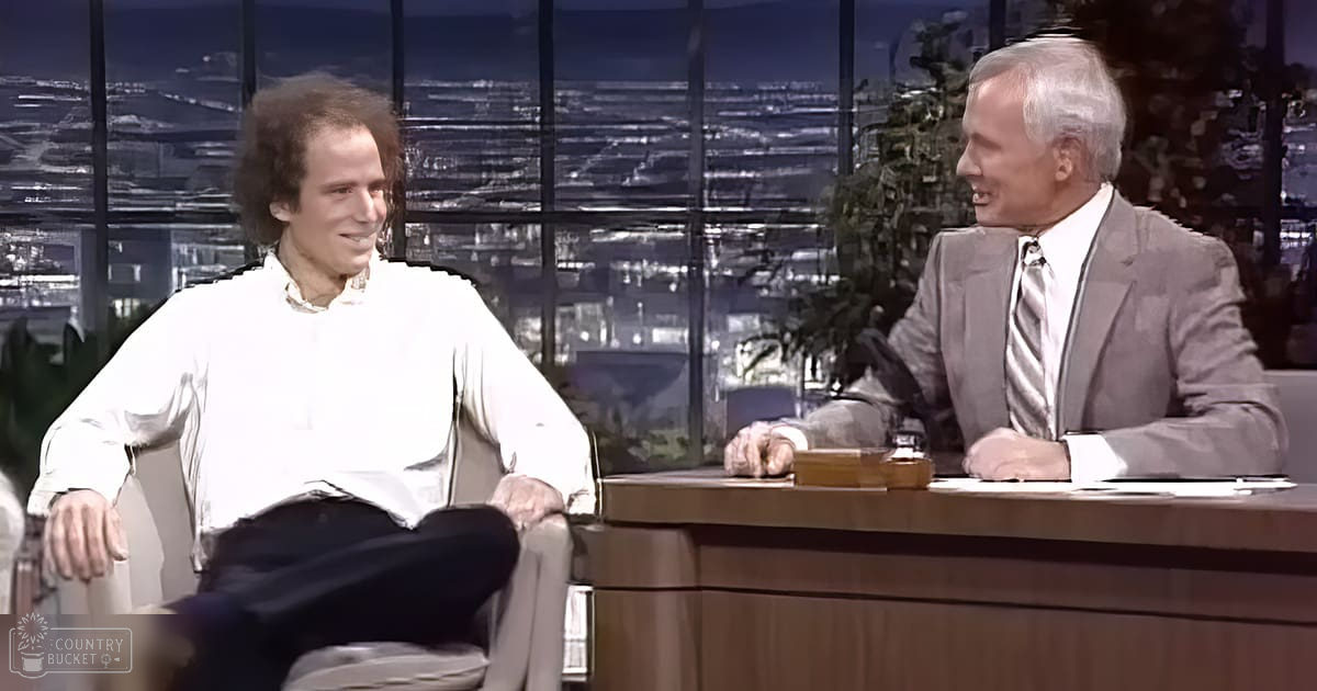 Adult male guest wearing white shirt and dark pants seated casually; older adult male host in gray suit jacket over white dress shirt with patterned tie, seated at wooden desk against large windows revealing a cityscape at night.