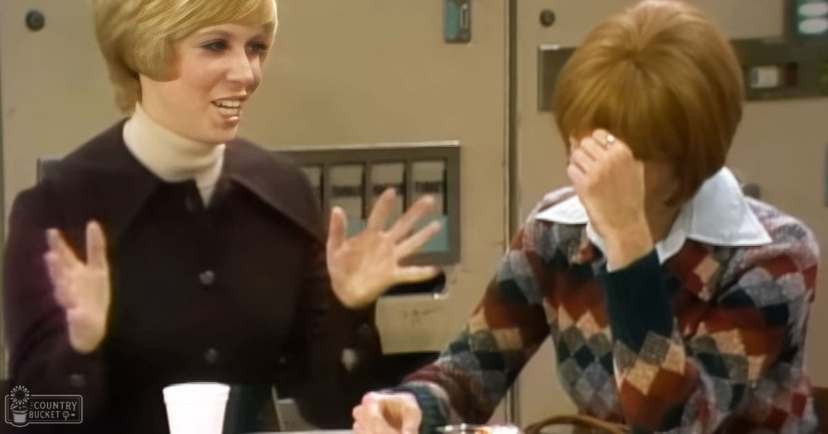 Two individuals in an office setting, one wearing a black double-breasted coat and white turtleneck, holding a disposable cup; the other in a plaid shirt with red, blue, green, and beige patterns, appearing contemplative. Beige walls suggest daytime lighting.