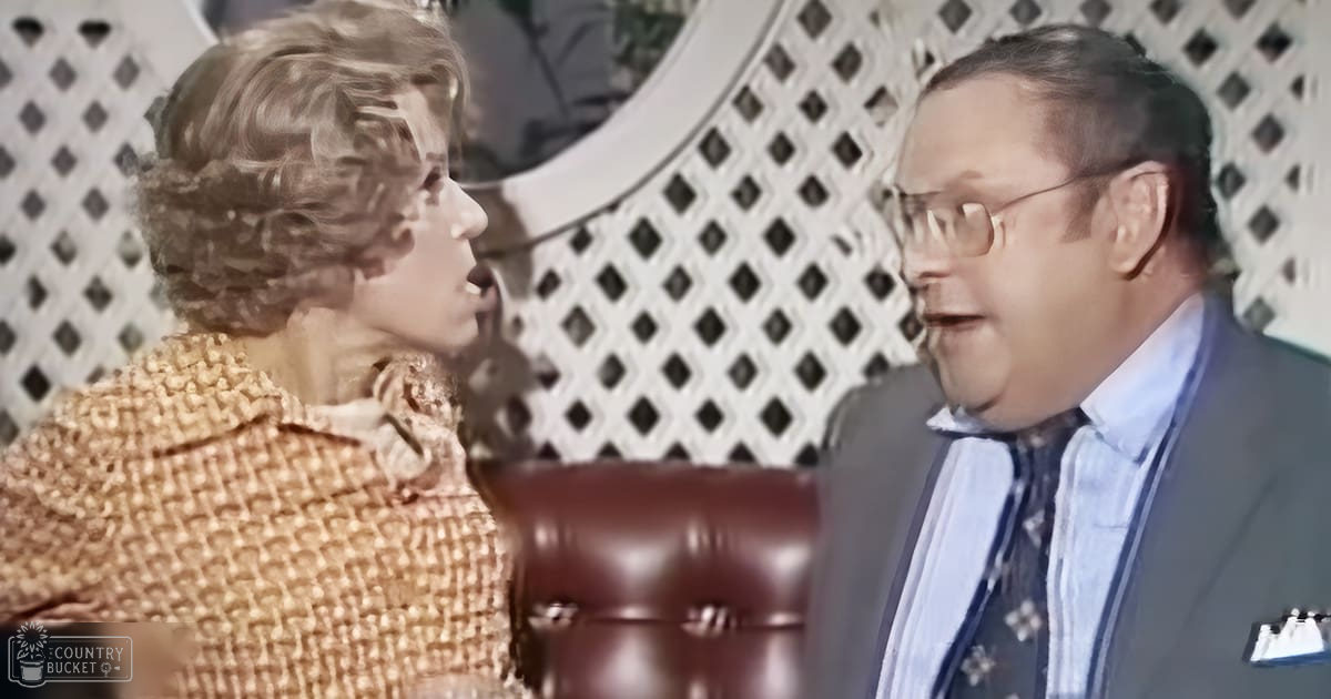 A middle-aged woman wearing a mid-20th-century tweed jacket with checkered patterns stands indoors, near an adult male in formal attire; background includes a lattice-patterned wall and circular mirror.
