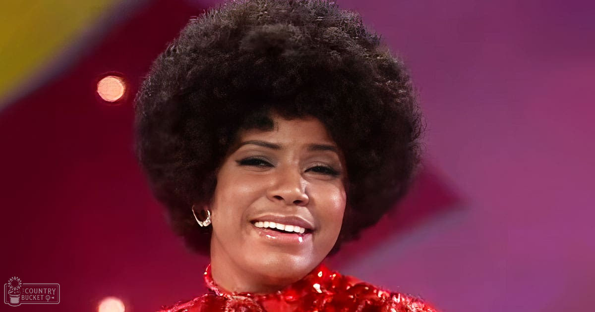 An adult female performer in a red sequined 1970s-style top with high neckline and short sleeves, adorned with earrings, on an indoor stage setting with warm lighting and purple hues.