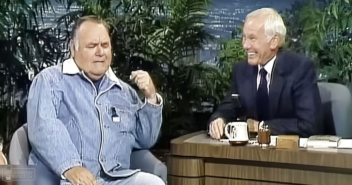 A middle-aged man in a denim jacket interviews an older adult male host dressed in a suit, seated at a wooden desk with decorative plants and urban backdrop in what appears to be a television studio setting.