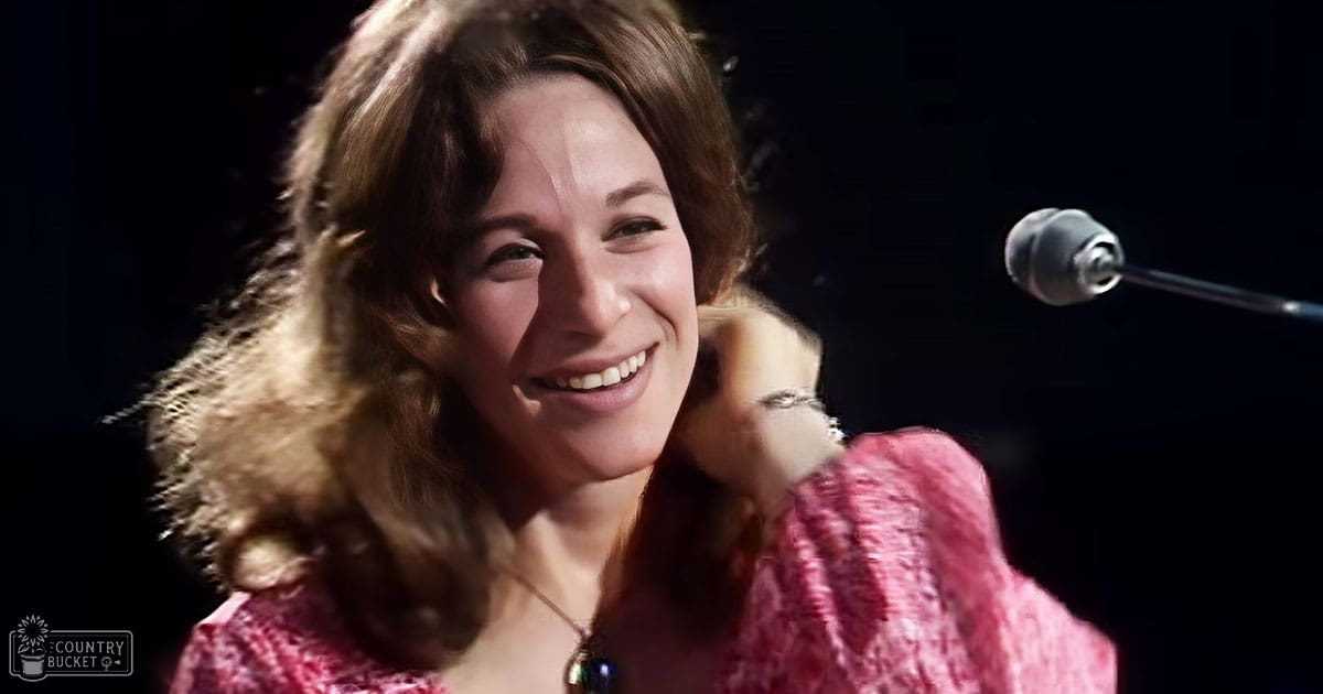 A young adult female in a relaxed, loose-fitting long-sleeved top with a floral or abstract pattern, standing on stage near a microphone and pop filter, wearing a necklace.