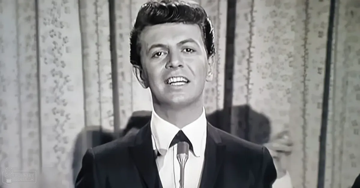 A young adult male wearing a tailored black suit with a white shirt and black tie stands at a microphone stand, suggesting a performance or recording session indoors.