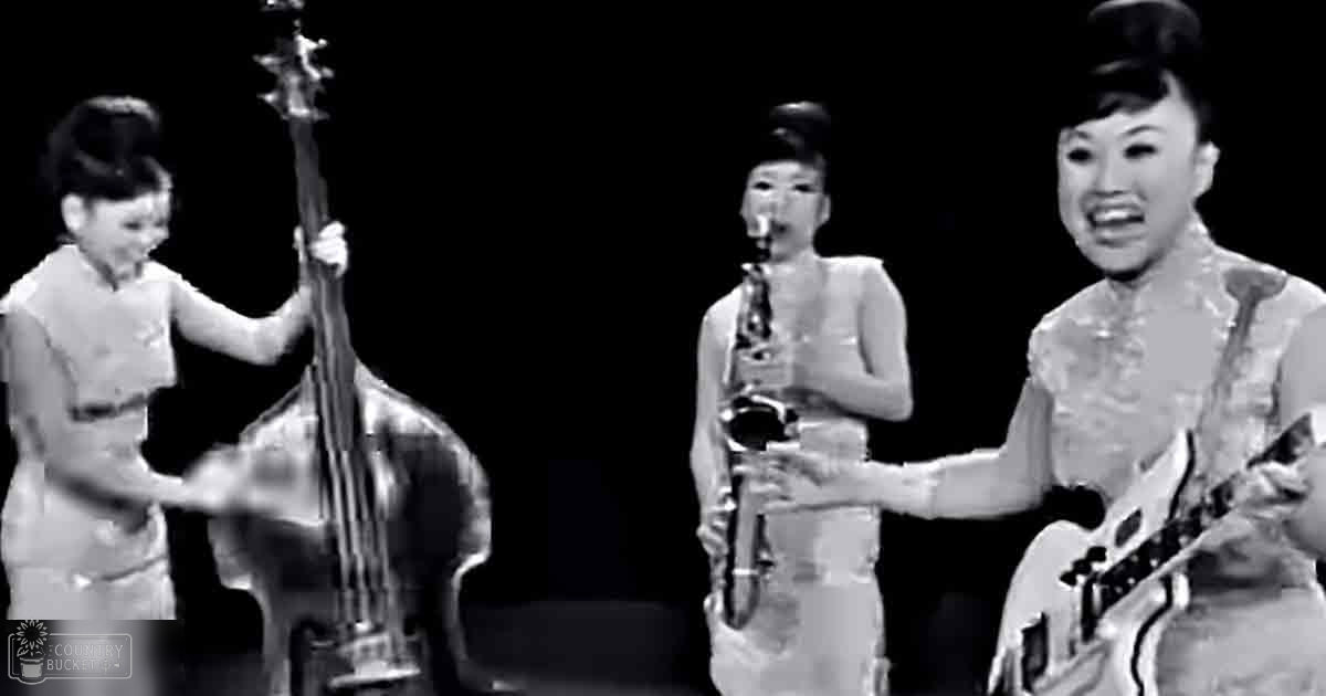 A musical performance on a stage under focused lighting, with young adult females in white dresses and holding double basses and saxophones.