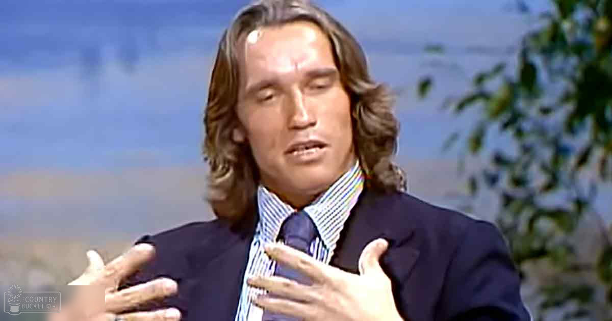 Adult male in a formal interview setting with a dark blue striped shirt and dark suit jacket against a backdrop of blurred greenery and blue sky, suggesting an outdoor filming location.