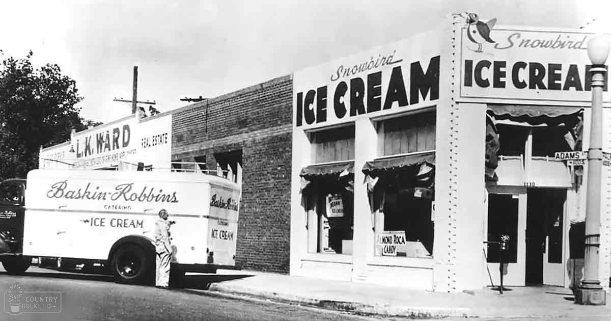 A mid-20th-century Baskin-Robbins ice cream truck parked in front of a 'Snowbird Ice Cream' shop with a brick facade and streetlamp, set against an urban backdrop.