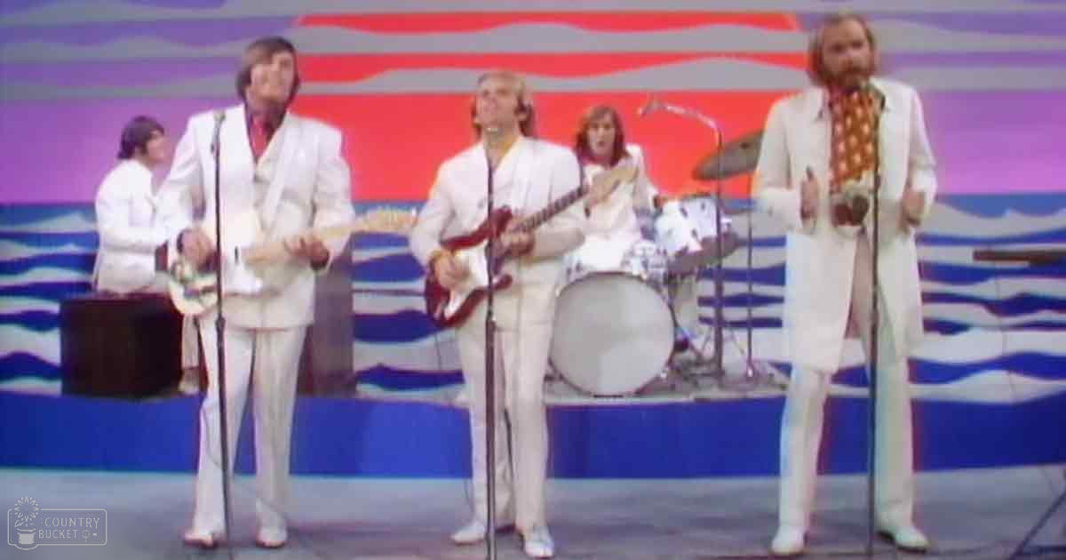 A vibrant stage set with bold red, white, blue, and purple abstract designs or waves backdrop, with a performance group in 1970s-style tailored suits.
