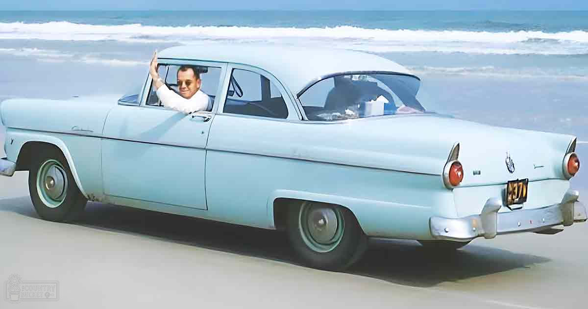 An adult male wearing a white shirt with rolled-up sleeves, sunglasses, and possibly a light-colored tie or scarf waves while seated inside an old blue vintage car parked on a sandy beach under natural daylight.