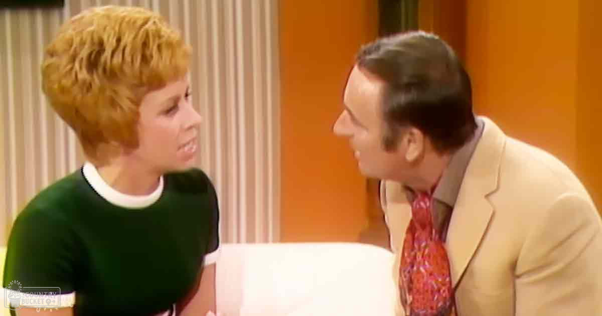 An adult female with short blonde hair, wearing a green T-shirt indoors near an adult male in a light beige suit jacket and red patterned tie, surrounded by striped curtains and an orange wall in the living room.