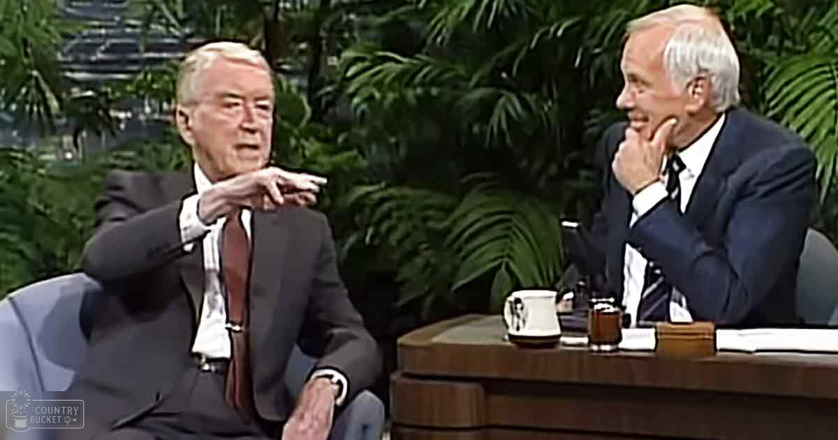 Indoor talk show set with older adult males wearing formal suits, lush greenery suggesting a tropical theme, and artificial lighting evenly illuminating the scene.