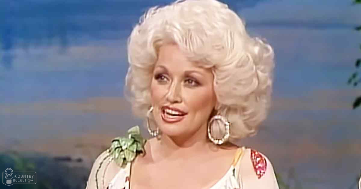An adult female performer wearing a loose, sleeveless white top with red and yellow floral patterns, accessorized with large hoop earrings and bracelet, set against an outdoor backdrop with greenery and blue sky.