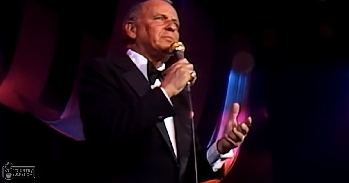 An older adult male performer in a black tuxedo holding a microphone, likely mid-20th century formal attire, performing on stage with dark purple and blue lighting behind him.