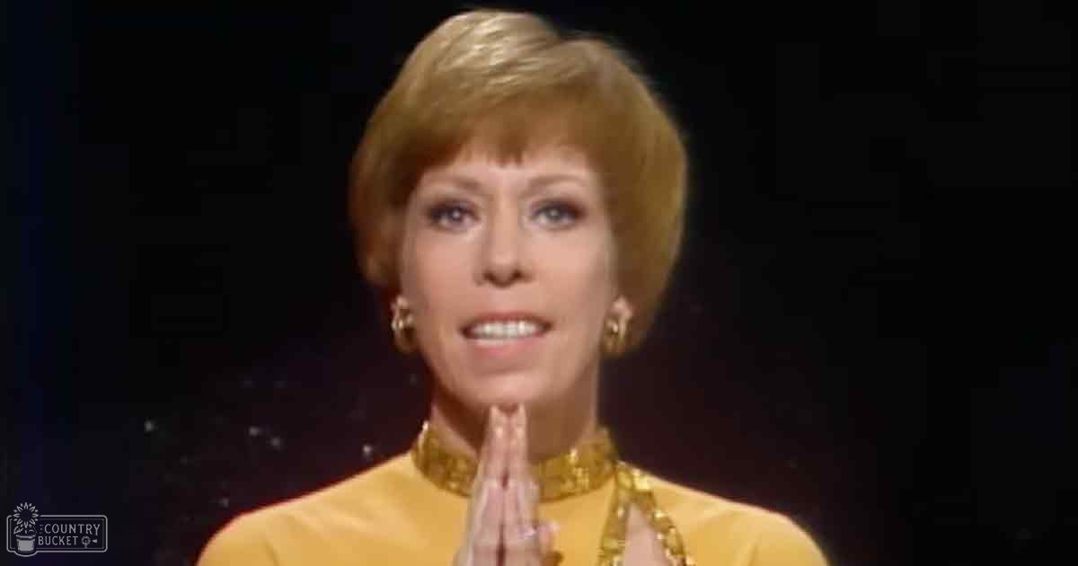 A middle-aged woman in an indoor setting, wearing a yellow, high-collared, intricately detailed formal garment with gold earrings and possibly a necklace. The background has star-like specks suggesting a space theme.