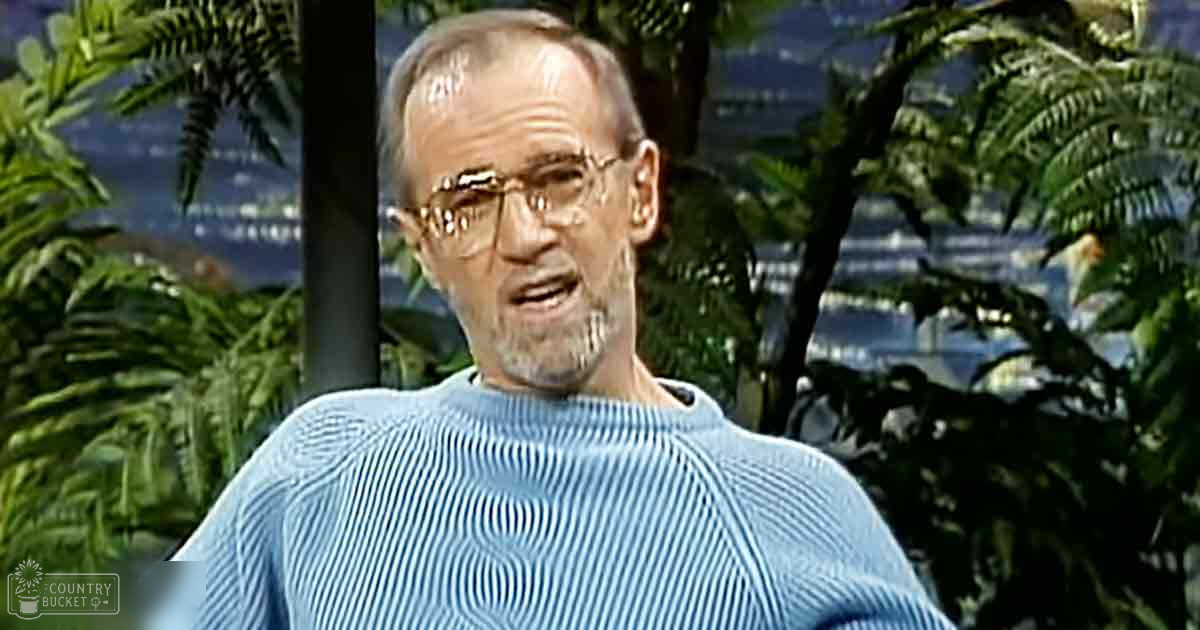 An older adult male in a light blue, ribbed sweater stands amidst lush tropical foliage under soft daylight.