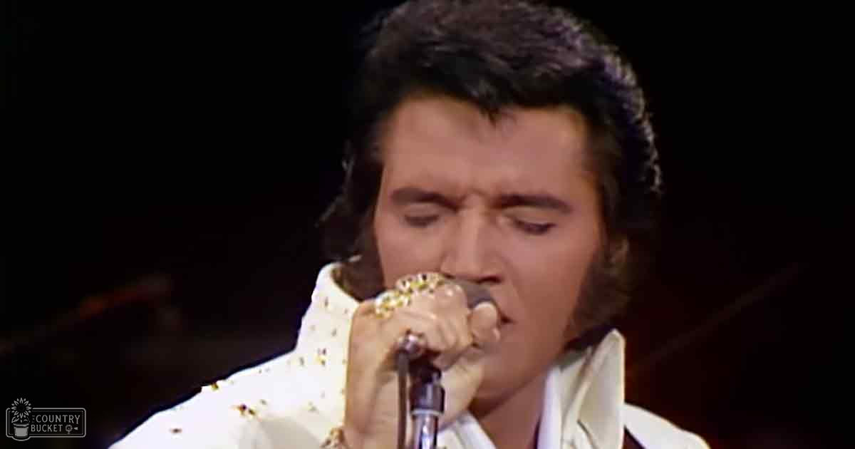 An adult male performer wearing a white stage costume with gold sequins, black trim, and holding a microphone on an indoor stage.