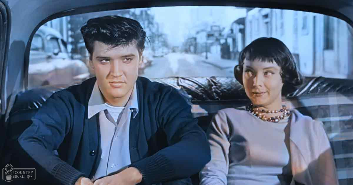A young adult male and female passenger are seated inside a mid-20th-century styled car in an urban setting during what appears to be daytime or early evening.