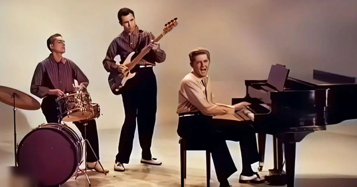 A musical performance scene with young adult male musicians in an indoor studio setting, with a grand piano and drum set components visible.