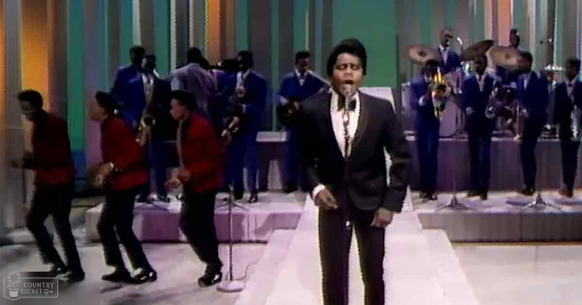 An adult male singer wearing a black suit, white shirt, and bow tie stands on stage with brass instruments being played by musicians behind him under professional stage lighting at an indoor venue.