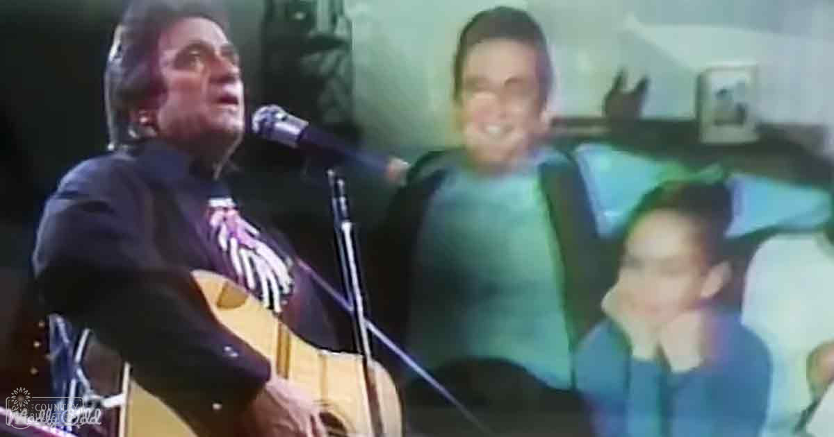 A performer singing into a microphone while holding an acoustic guitar, wearing a dark-colored jacket with visible graphic design, surrounded by two casually dressed individuals in a residential indoor setting with natural light and neutral tones.