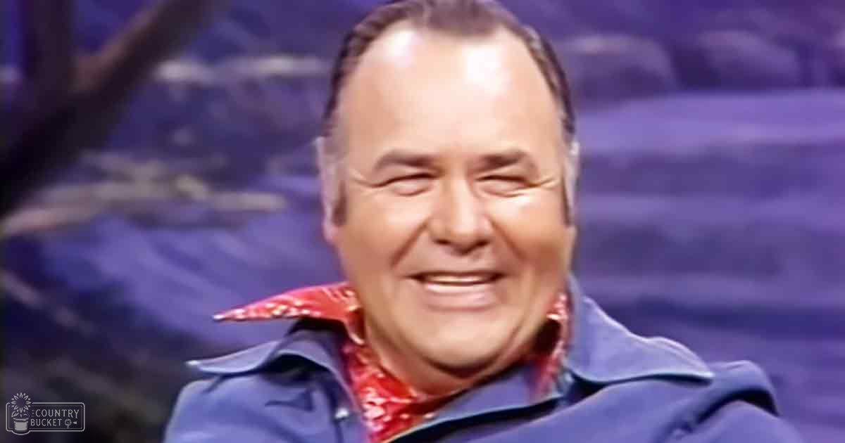 A middle-aged man wearing a blue jacket and red bandana on stage, suggesting a past era performance setting with blurred lights in the background.