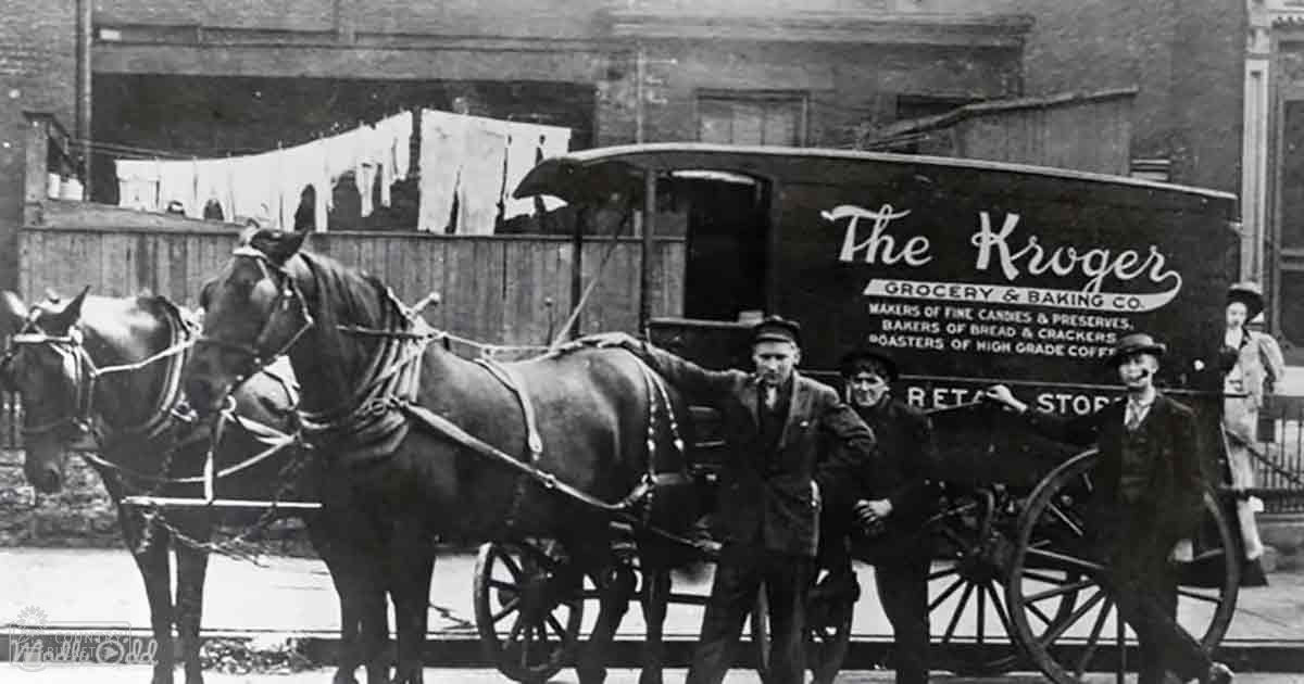 Two dark brown draft horses pulling a delivery wagon labeled 'The Kroger Grocery & Baking Co.' in an urban setting with brick buildings and hanging laundry, early 20th century scene.