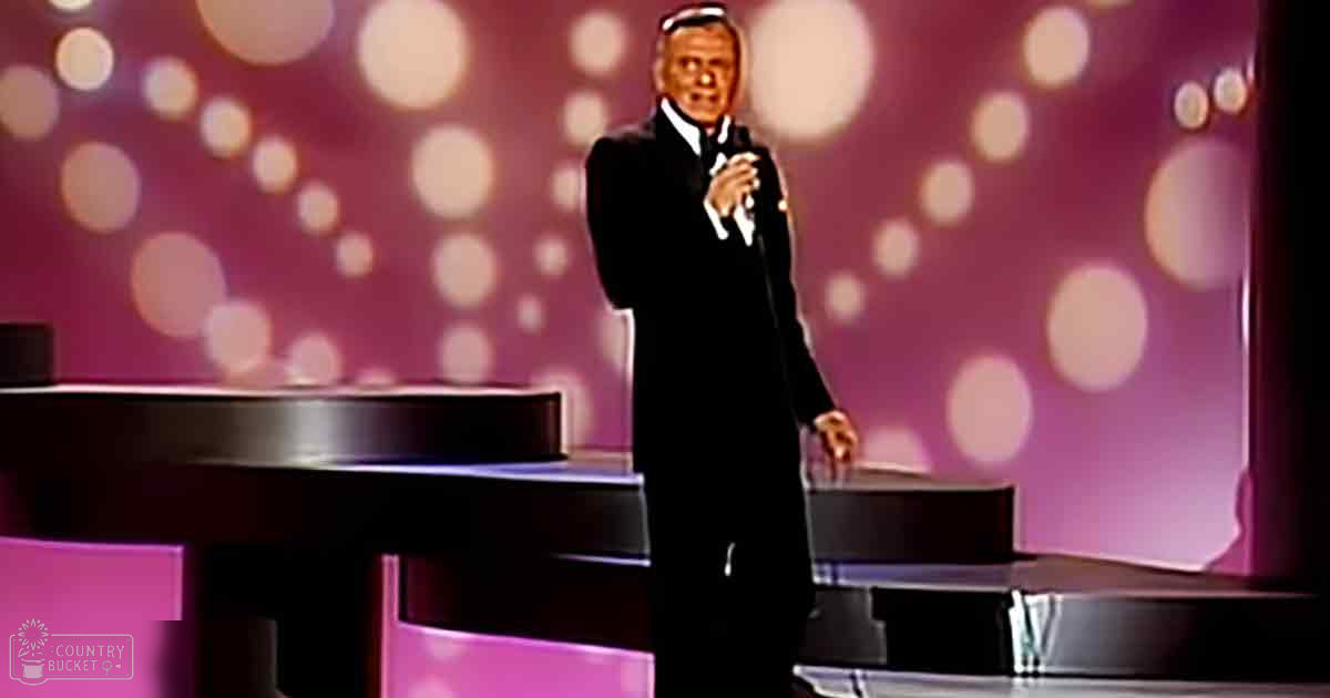An older adult male performer in a formal black suit at an awards ceremony stage with bokeh lights and pinkish backdrop, text 'Madly Odd' visible.