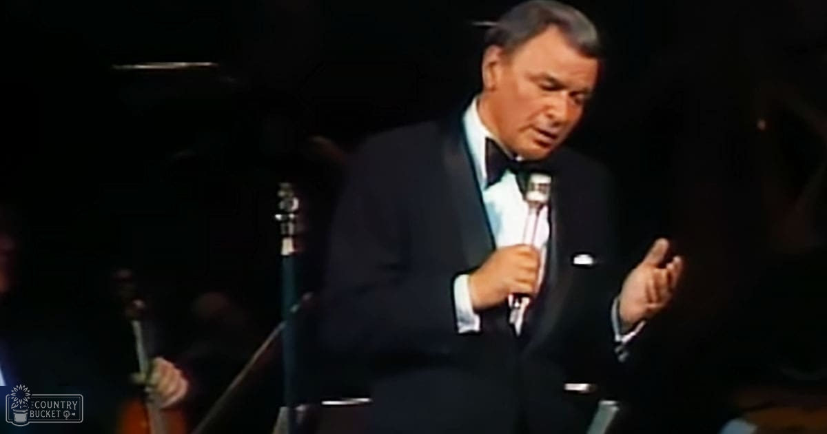 An older adult male singer in a mid-20th century formal attire, possibly on stage with an orchestra setup behind him.